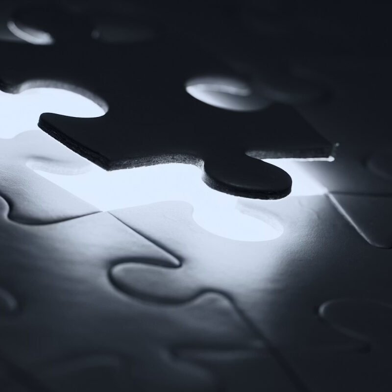 A dark puzzle piece hovering over a glowing gap in a mostly dim jigsaw.