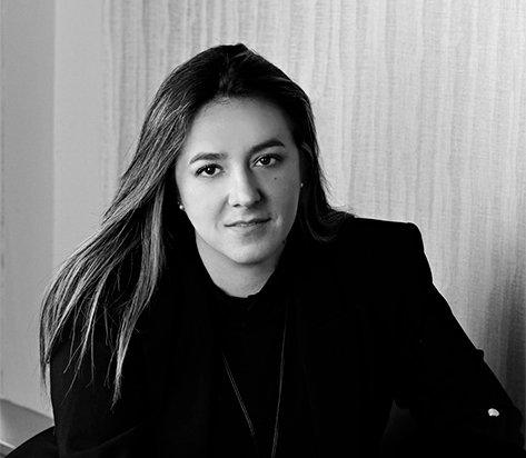 Portrait of a woman with long hair, wearing a dark blazer, looking at the camera in a neutral setting.