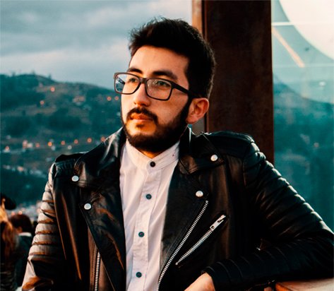 Man with dark hair and beard in a black leather jacket and white shirt, looking off-camera outdoors.