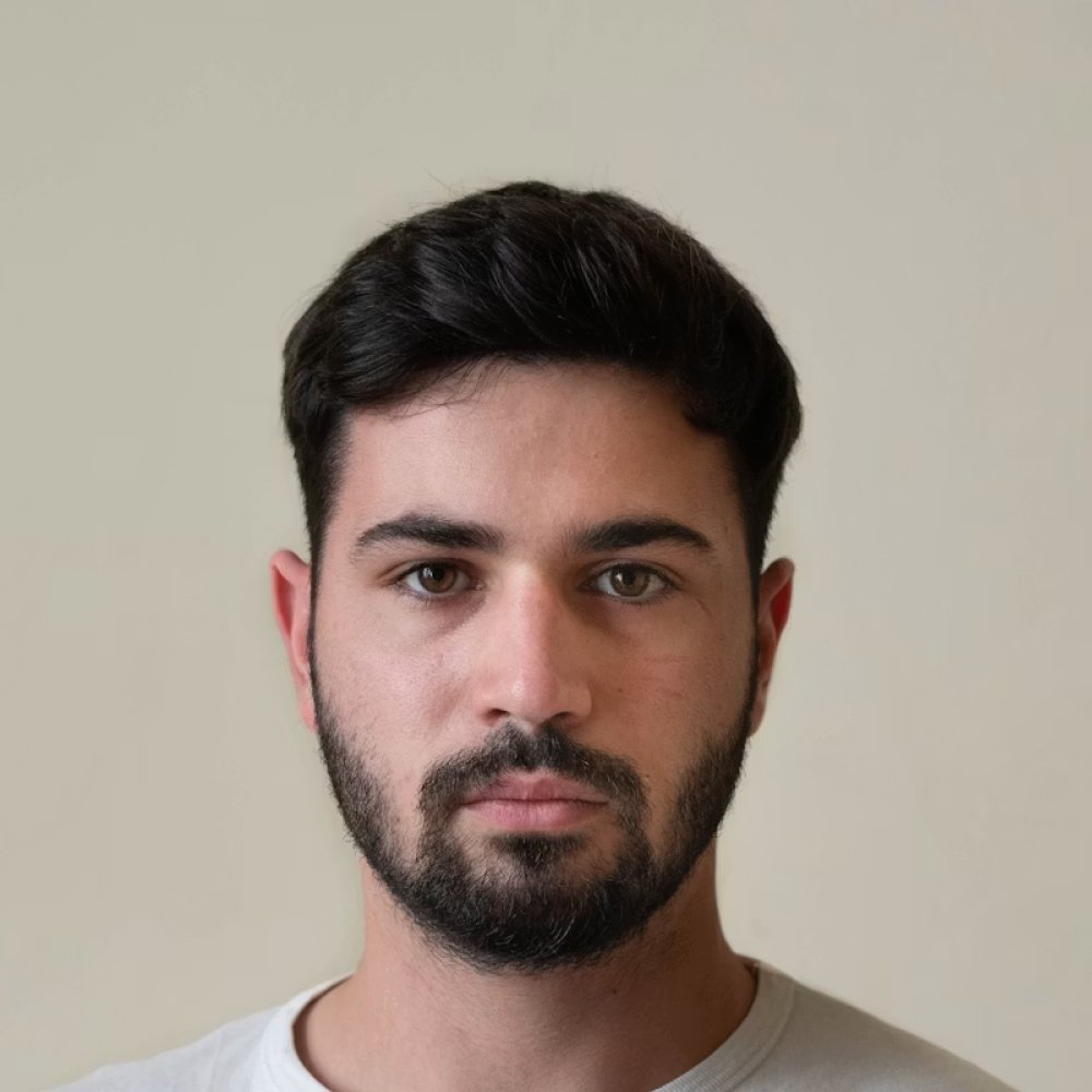 Close-up portrait of a man with short dark hair and a trimmed beard looking at the camera on a neutral background.